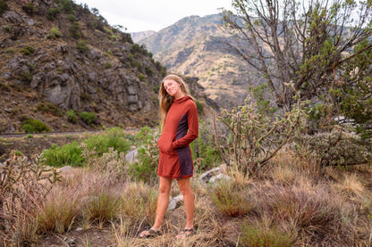 A woman stands outdoors in a rocky, brushy mountain landscape wearing a long-sleeve athletic dress in rust orange and dark gray. The dress has a kangaroo pocket and is styled with dark sandals.