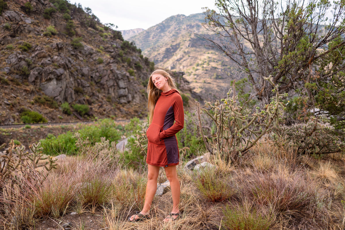 A woman stands outdoors in a rocky, brushy mountain landscape wearing a long-sleeve athletic dress in rust orange and dark gray. The dress has a kangaroo pocket and is styled with dark sandals.