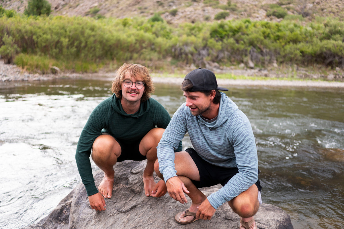 Two men crouching on a rock by a river, wearing long-sleeve athletic shirts and shorts in an outdoor natural setting.